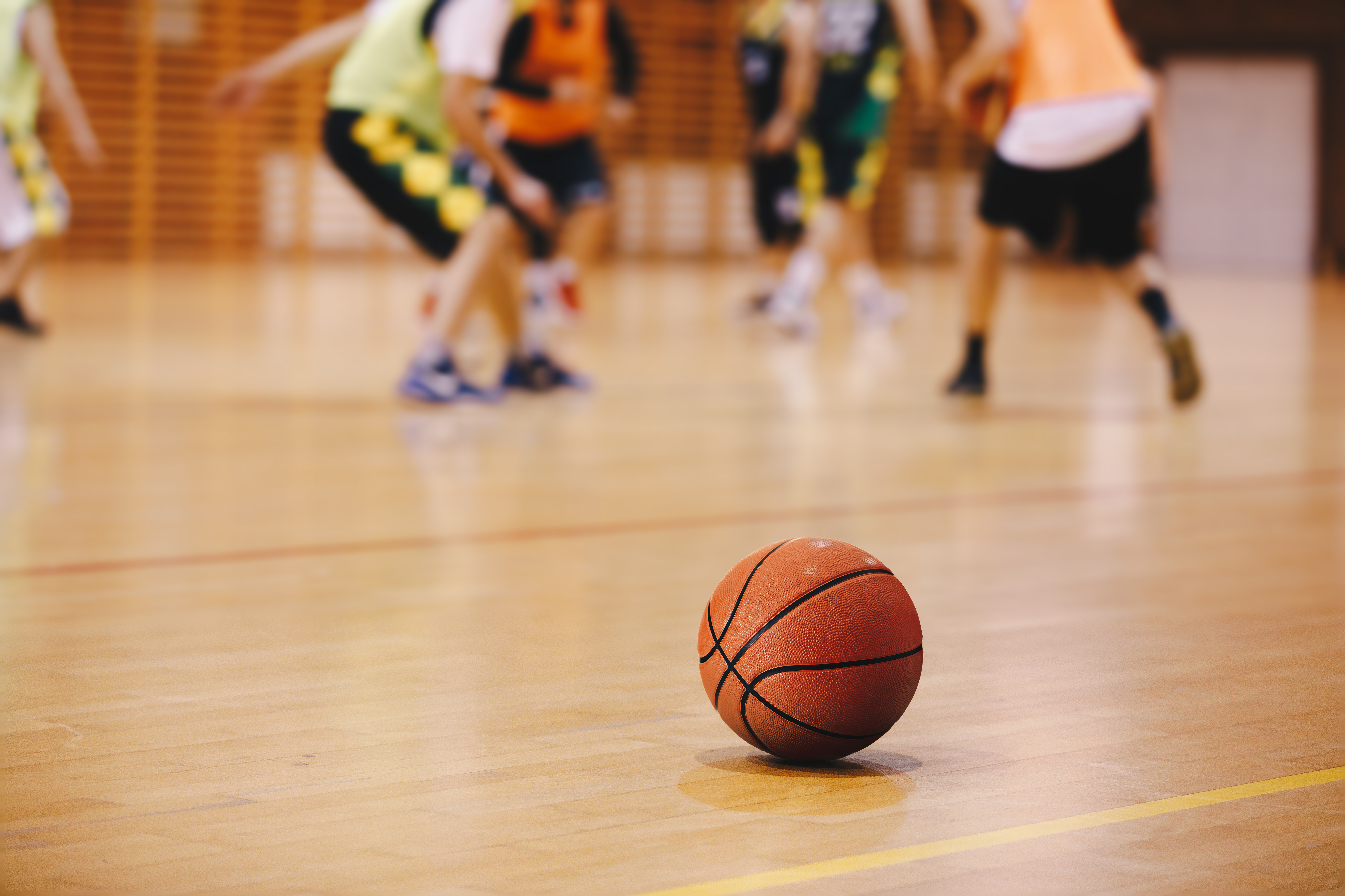 Basketball on Wooden Court Floor Close Up with Blurred Players Playing Basketball Game in the Background.